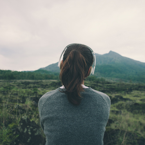Image of woman with headphones on, overlooking grassy fields leading to a mountain in the distance.