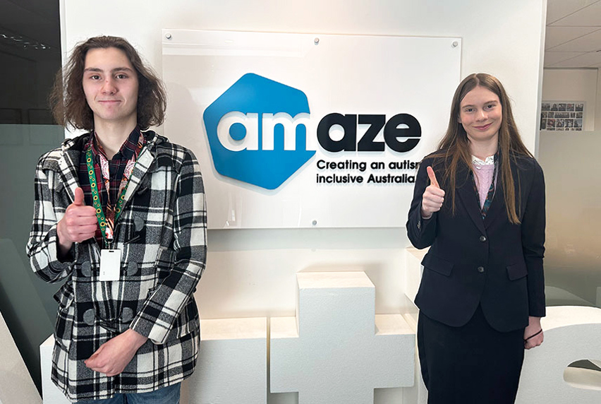 Two individuals pose together in front of a large, illuminated "amaze" sign, smiling and giving a thumbs up.