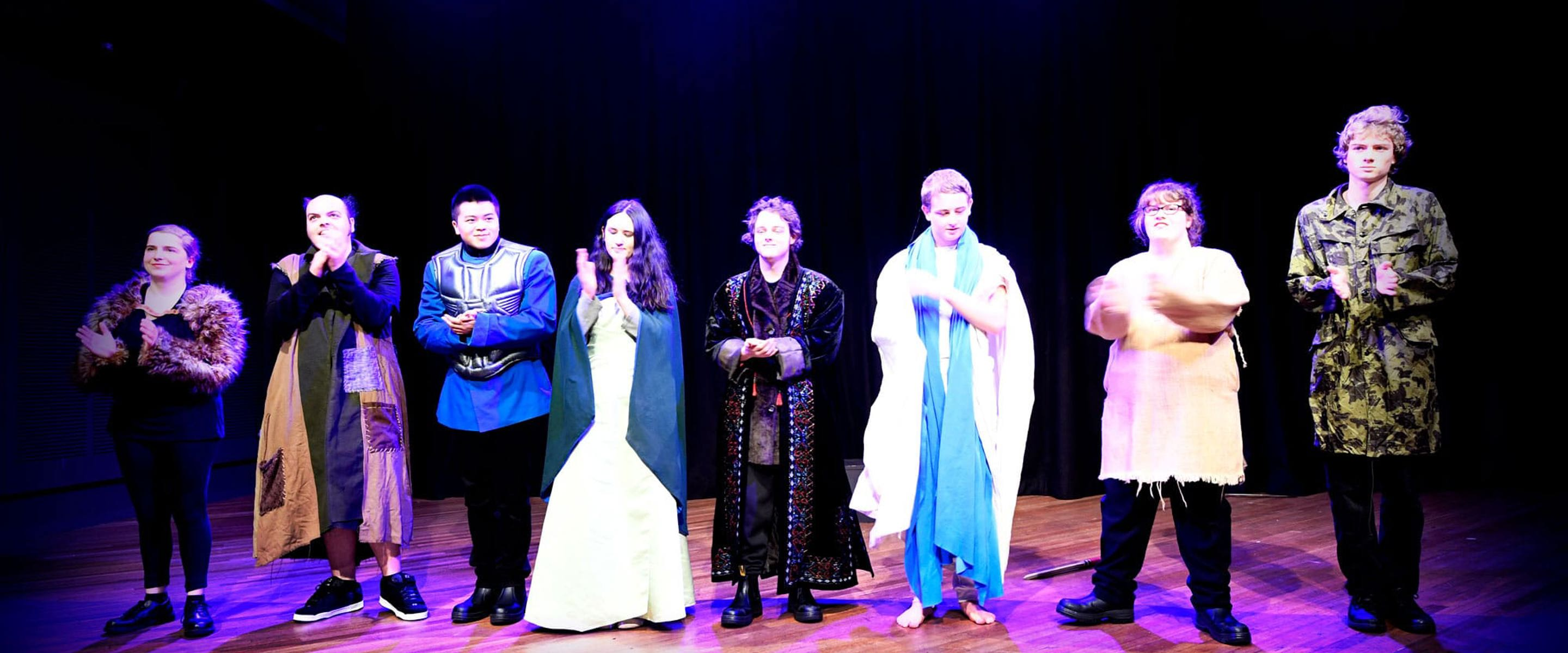 A group of people standing on a stage under theatre lights with costumes on