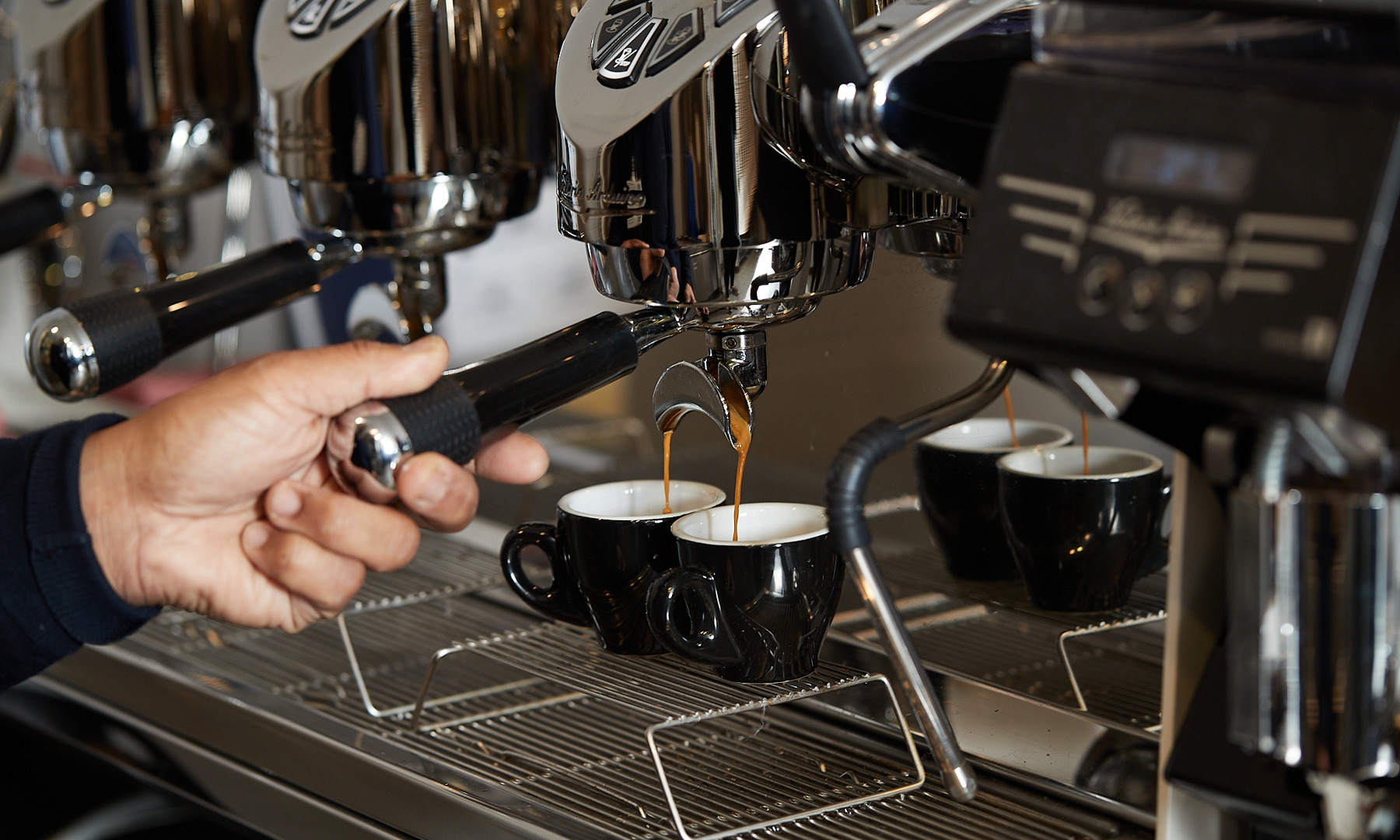 Close-up image of a barista's hand holding the portafilter of a coffee machine as the machine extracts coffee into two small mugs.