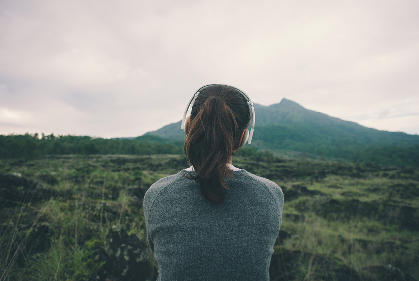 Image of woman with headphones on, overlooking grassy fields leading to a mountain in the distance.