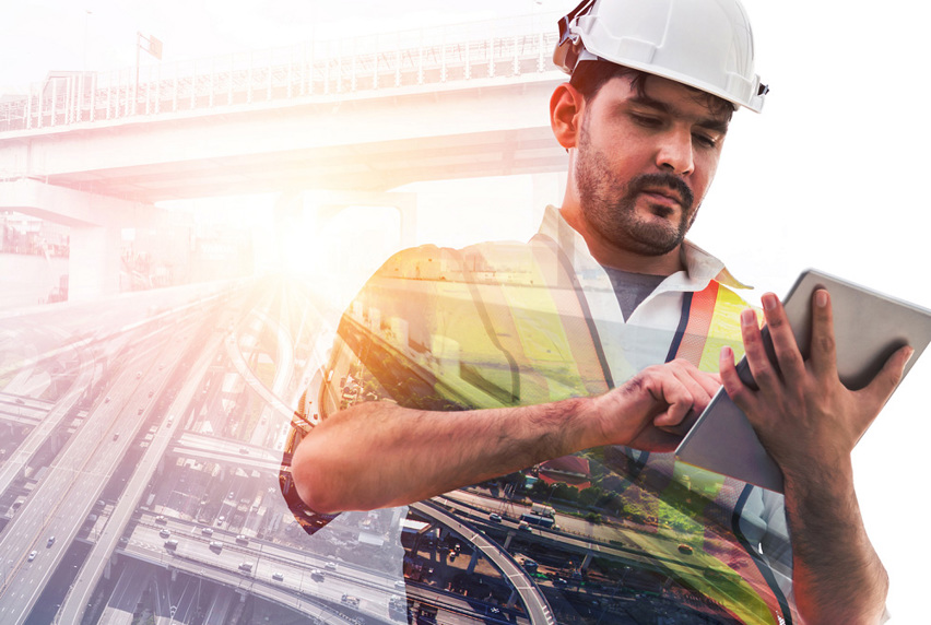 Man with construction hat, hi-vis vest, working on a tablet device, with an image of freeways and a bridge in background