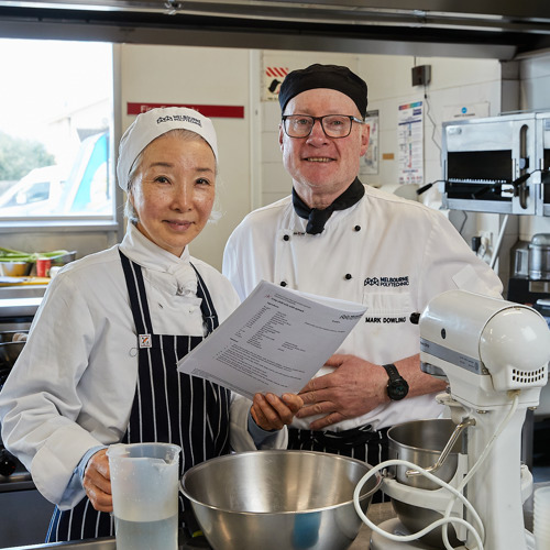 A man and woman in Melbourne Polytechnic Cookery uniforms holding papers in a commercial kitchen.