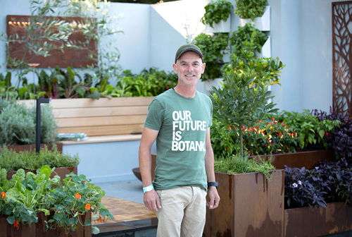 Justin Rhodes in a green t-shirt standing in front of their garden design, surrounded by colorful flowers and lush greenery.