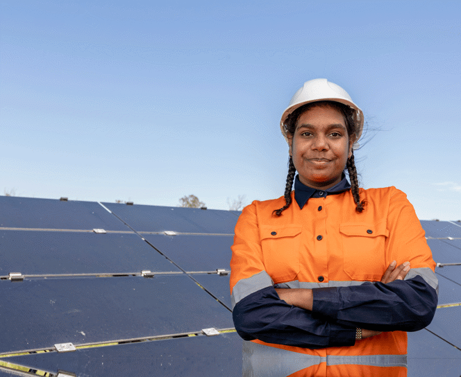 Smiling woman standing outside in safety work clothing and helmet with her arms crossed in front of solar panels.