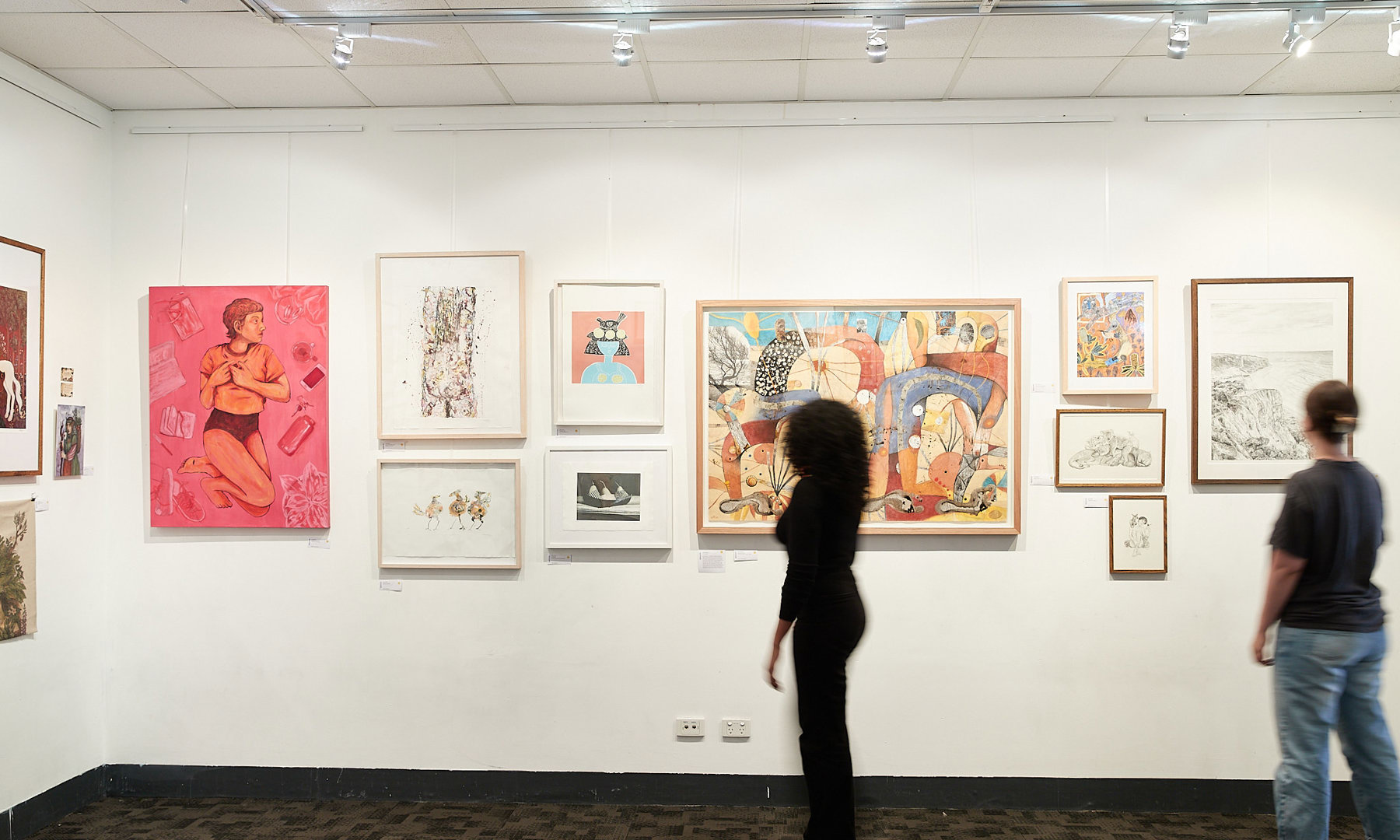 A gallery wall with student illustration artwork displayed. There are two visitors observing the art work.