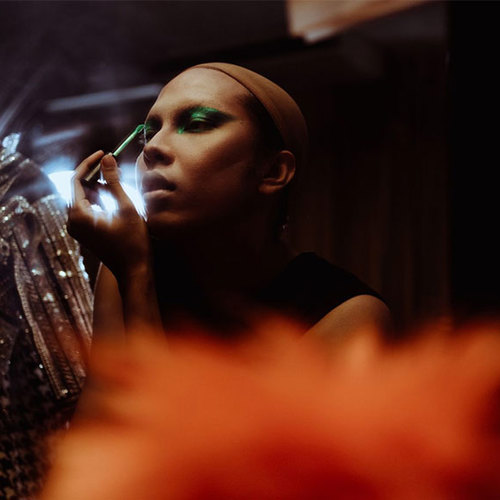 In mood lighting, a theatre actress applies green eyeshadow to her eyelids, while looking in a mirror.