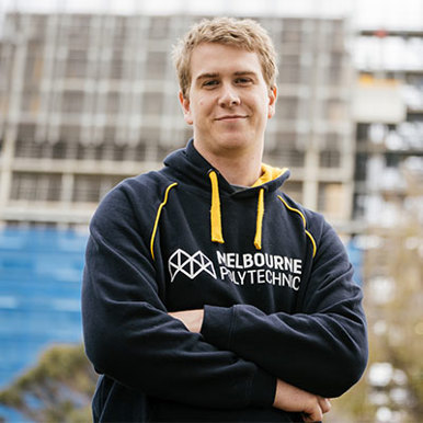Image of a young blonde male wearing a blue Melbourne Polytechnic jumper smiling. He is standing in front of a building. 