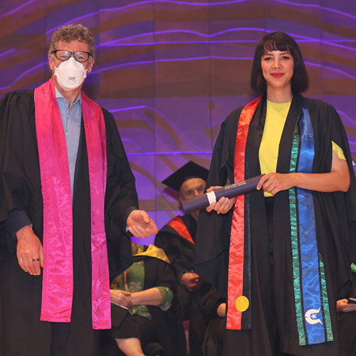 A woman in her graduation gown stands next to a man in a mask, capturing a moment of accomplishment and awareness at her graduation