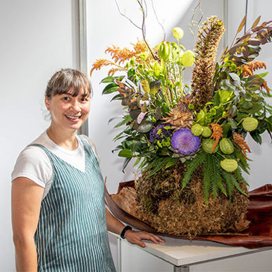 A  brunette woman stands beside a sizable flower arrangement smiling
