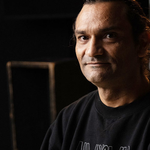  Image of a man in a black shirt with medium length hair, staring down the camera in mood lighting  