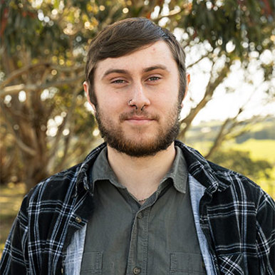 A bearded young man wearing a flannel shirt with trees in the background