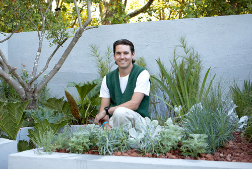 Jamie Greentree's 'Permeable Paradise' garden: Jamie, wearing a green vest and white t-shirt, crouches amidst lush plants and a neatly arranged garden bed in his design.
