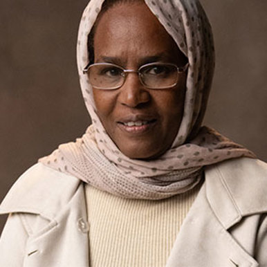 A woman wearing a light pink head scarf with glasses and a beige jacket