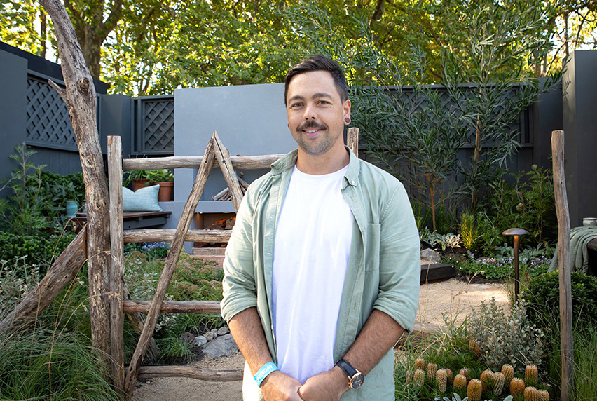 Leigh Hudson stands in a garden that features wooden decor, banksias, and other native plants. He wears a white t-shirt beneath a pale green shirt.
