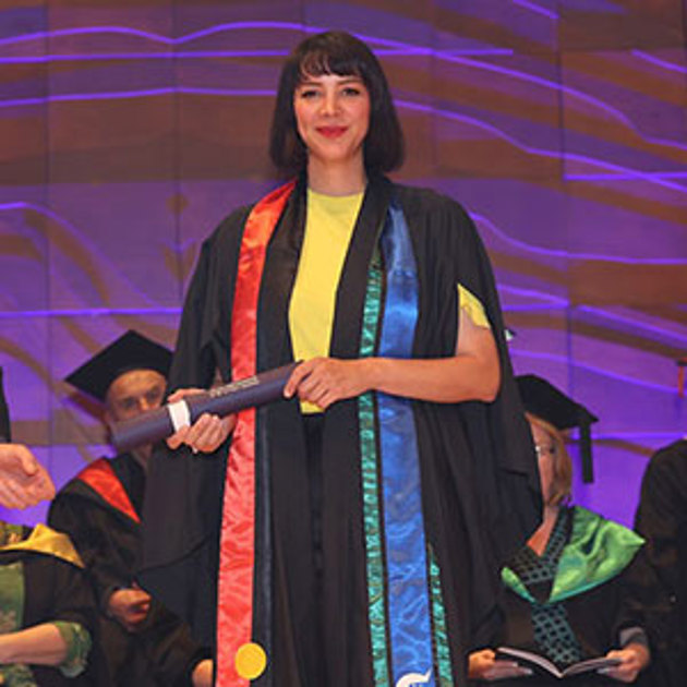 A woman with medium-length black hair holding a testamur cylinder wearing a graduation gown and an Australian Aboriginal and Torres Strait Islander flag stole.