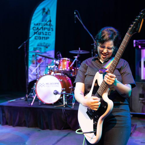 A passionate guitar player on stage at a music competition, joyfully performing with a white electric guitar, with a drummer and another guitarist in the background, under vibrant stage lighting.