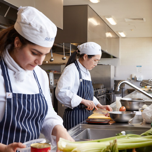 Two hospitality students, in white cook's shirts and navy and white striped aprons, don Melbourne Polytechnic cook's caps while engaged in the process of food preparation.